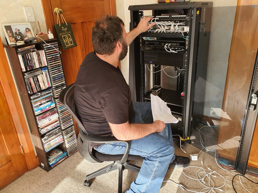 Richard Barlow, a bearded gentleman in jeans and T-shirt, sits in front of a newly-furnished larger network cabinet.